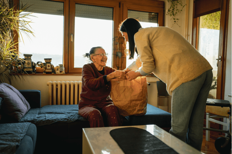 A woman hands a paper bag to an elderly woman sitting on a couch in a cozy living room, highlighting the warmth of the home care ecosystem.