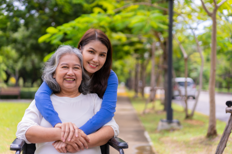 A woman embraces an elderly woman seated in a wheelchair on a tree-lined path, their smiles reflecting the joy of Colorado home care.