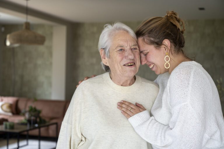 Elderly woman smiling warmly as a young woman hugs her and laughs in a cozy home environment.